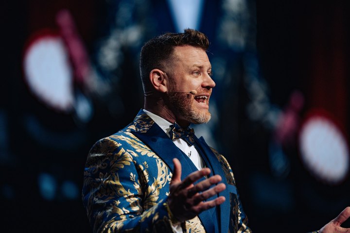 A speaker in a blue and gold jacket speaking with an open hand gesture
