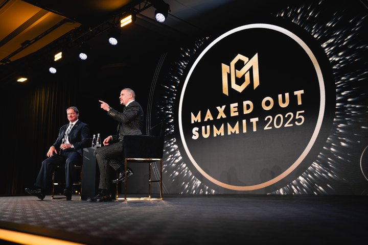 Two speakers in a stage conversation beside the Maxed Out Summit 2025 screen