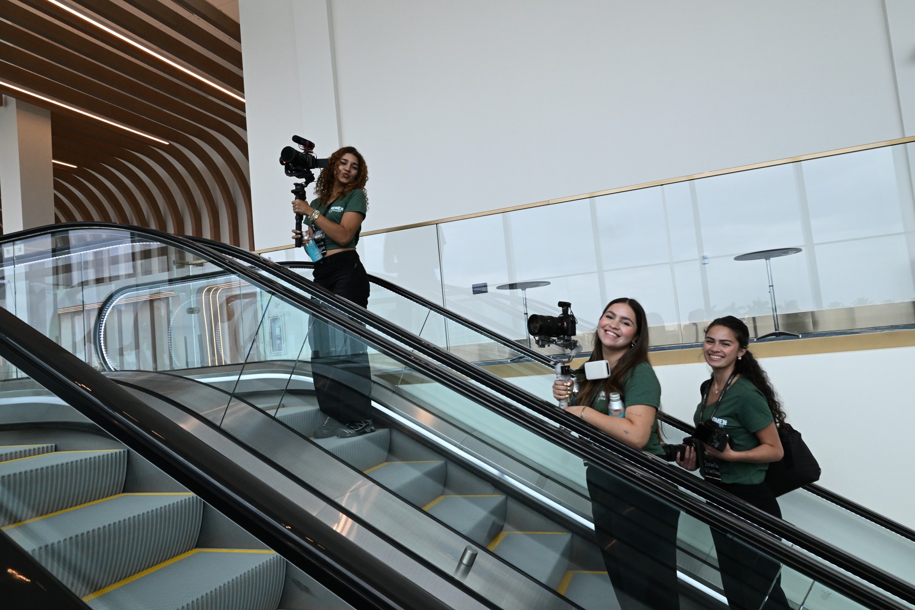 Three smiling crew members carrying cameras on an escalator at Women and Wealth
